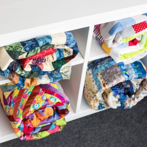 84472177 - patchwork, sewing and fashion concept - colorful finished quilts in the studio at white shelves with few storage compartments, the warehouse of finished products, top view